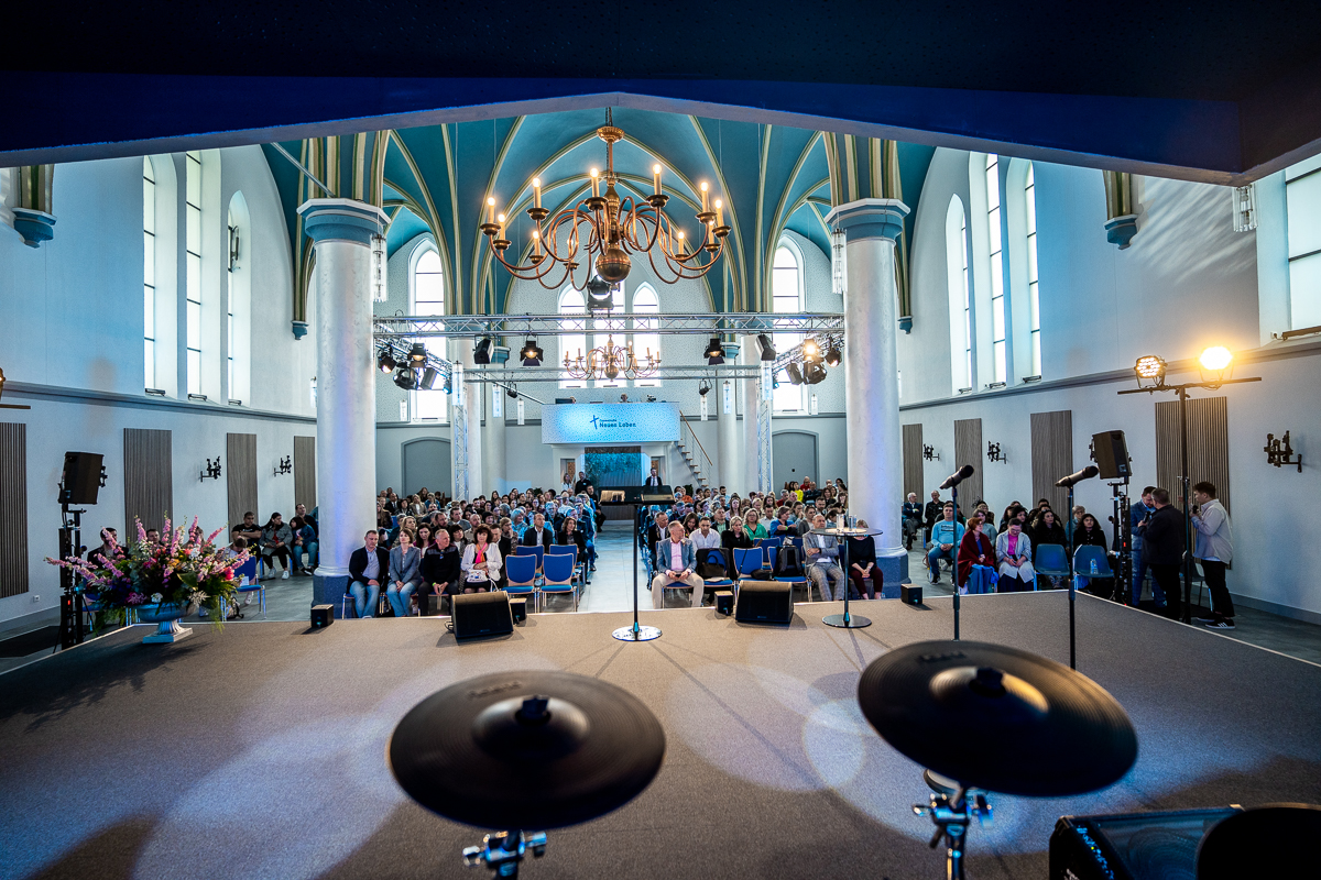 Eine Gruppe von Menschen versammelt sich in einem hellen Kirchengebäude in Aachen. Sie sitzen in Bänken und hören der Predigt des Pastors zu. Das Licht strömt durch die farbigen Fenster, während eine Atmosphäre der Ruhe und des Glaubens herrscht.