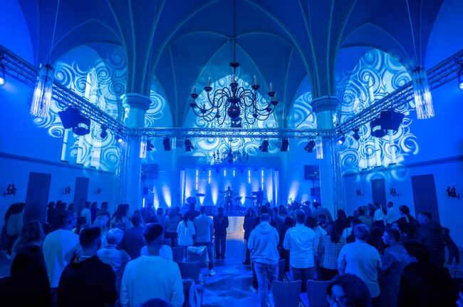 Mitglieder der Gemeinde Neues Leben Kirche in Aachen arbeiten an der Steuerung von Licht und Ton während eines Gottesdienstes. Sie konzentrieren sich darauf, eine einladende und inspirierende Atmosphäre zu schaffen.
