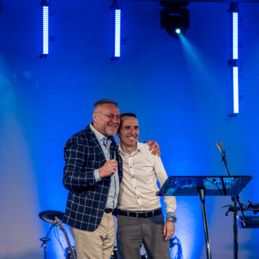 Mitglieder der internationalen Gemeinde Neues Leben in Aachen lächeln einander zu und umarmen sich herzlich. Die Atmosphäre ist geprägt von Freundschaft und Zusammenhalt.